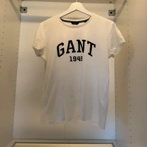 Vit GANT t-shirt med tryck - Vit t-shirt från GANT med svart tryck 'GANT 1949' på bröstet. Klassisk rund hals och korta ärmar. Tillverkad i mjuk bomull för en skön och avslappnad känsla. Perfekt för en clean och sportig look.