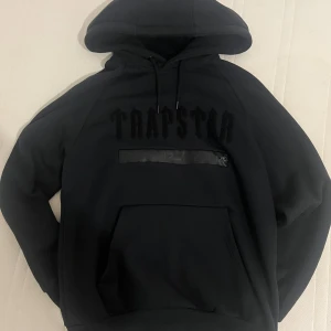 Svart Trapstar hoodie med brodyr - Priset kan diskuteras!Svart hoodie från Trapstar med broderad logga på bröstet och en unik detalj med textband. Stor huva med broderad T på baksidan. Känguruficka framtill och snörning i halsen. Tillverkad i 80% bomull och 20% polyester för en skön och mjuk känsla.