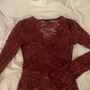 Vinröd spetsblus från Gina Tricot XS - Säljer en vinröd blus från Gina Tricot i storlek XS. Blusen är helgjord i genombruten spets med blommigt mönster, har lång ärm och v-ringad hals. Har ett litet hål i vänstra ärmhålan, använd ca 3 ggr