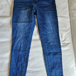 Blå high waist skinny jeans med bälten - Snygga blå jeans med hög midja och skinny fit. Byxorna har fyra framknappar och dubbla bälten med guldfärgade spännen. Klassisk femficksmodell och stretchigt denimtyg som sitter tajt och bekvämt. Perfekt för dig som gillar trendiga detaljer.