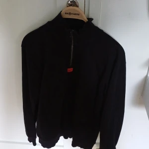 Svart half zip sweatshirt från HUGO BOSS - Svart sweatshirt från HUGO BOSS med half zip och hög krage. Tröjan har en stilren look med ribbade muddar och en liten röd detalj vid dragkedjan. Perfekt för dig som gillar minimalistisk stil och vill ha något som funkar året runt.