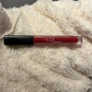 Huda Beauty Liquid Matte läppstift - Flytande matt läppstift från Huda Beauty i en djup röd nyans. Kommer i en stilren, frostad tub med svart lock. Perfekt för dig som vill ha intensiva, matta läppar med lång hållbarhet. Använt endast på handen 