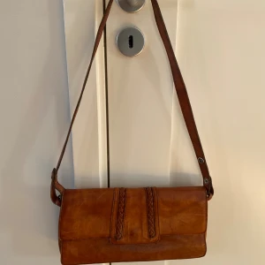 Brun axelväska i skinn med flätad detalj - Snygg brun axelväska i äkta skinn med justerbart axelband och flätade detaljer på framsidan. Väskan har flera fack med dragkedja och ett klassiskt metallspänne. Perfekt storlek för det viktigaste och ger en vintagevibe till din outfit. Mått Bredd: ca 31 cm Höjd: ca 18 cm  Kommer från ett djur och rökfritt hem.