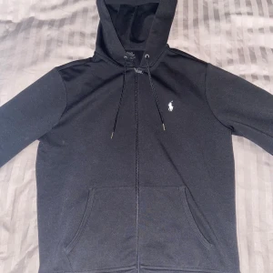 Svart hoodie från Polo Ralph Lauren - Svart hoodie från Polo Ralph Lauren med dragkedja, huva och snörning. Klassisk vit logga på bröstet och känguruficka framtill. Tillverkad i mjuk bomullsmix, perfekt för chill dagar eller när du vill ha en clean look.
