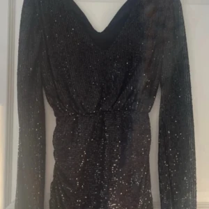 Svart glittrig klänning Meet Me There - Svart klänning från Meet Me There med långa ärmar och djup v-ringning. Klänningen är täckt av glittriga paljetter och har en markerad midja som ger en snygg siluett. Perfekt för dig som vill sticka ut med en festlig look.
