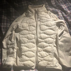 Beige cardigan Peak Performance M - Snygg beige pufferjacka från Peak Performance i storlek M. Jackan har quiltad framsida, dragkedja och hög krage. Materialet är syntet och polyester, perfekt för kyliga dagar. Stilren design med diskret logga på bröstet och långärmade ärmar.