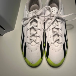 Adidas Crazyfast vita och gula fotbollsskor - Säljer ett par Adidas Crazyfast fotbollsskor i vitt med svarta ränder och neongula detaljer. Skorna har snörning, platt sula med dobbar och är tillverkade i syntetmaterial. Perfekta för dig som vill sticka ut på planen med stilrena och sportiga skor.