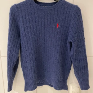 Mörkblå stickad tröja Polo Ralph Lauren - Mörkblå stickad tröja från Polo Ralph Lauren med klassiskt kabelstickat mönster och röd broderad logga på bröstet. Rund halsringning och långa ärmar. Perfekt för lager-på-lager under höst och vinter. Den är lite nopprig, det är pga av det fluffiga, luddiga och stickade materialet, går att åtgärda på cirka 5-10 min! Axelbredd 37 och 58 längd 