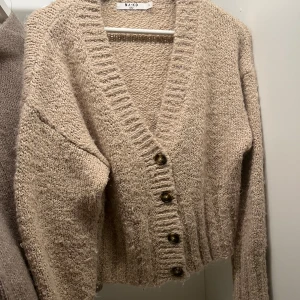 Beige stickad kofta från NA-KD - Mysig beige stickad kofta från NA-KD med djup v-ringning och stora knappar framtill. Koftan har långärmad design och ribbade muddar vid ärmslut och nederkant. Perfekt för lager-på-lager och chill dagar.