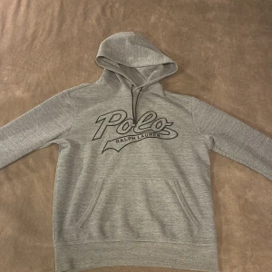 Grå hoodie från Polo Ralph Lauren - Snygg grå hoodie från Polo Ralph Lauren med stor svart logga framtill. Tröjan har huva med snörning och en klassisk känguruficka. Mjuk och skön bomullsblandning, perfekt för chill dagar. Långärmad och normal passform.