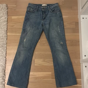 Blå bootcut jeans med blommigt mönster - Snygga blå jeans med bootcut passform och blommigt mönster på låret. Jeansen har klassisk femficksdesign, dragkedja och knapp framtill.