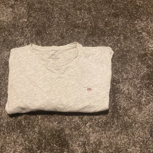 Ljusgrå t-shirt från GANT - Snygg ljusgrå t-shirt från GANT med klassisk passform och rund hals. T-shirten har diskret GANT-logga med sköld på bröstet och är tillverkad i mjuk bomull. Perfekt basic-plagg med korta ärmar och stilren look.
