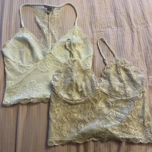 Två spetsiga linnetoppar i beige och vitt - Säljer två snygga linnetoppar i spets, en i vit och en i beige. Båda har tunna axelband, v-ringad hals och blommigt spetsmönster. Den vita är från New Yorker och den beige ifrån BikBok