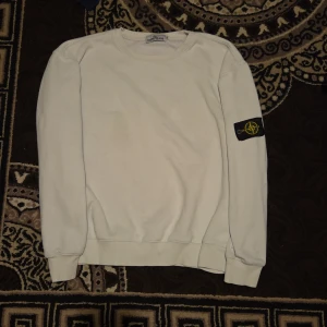 Vit Stone Island sweatshirt XL - Vit sweatshirt från Stone Island i storlek XL. Klassisk rund halsringning och ribbade muddar. Ikonisk svart patch med gul logga på vänster ärm. Tillverkad i mjuk bomullsblandning för skön känsla. Perfekt för dig som gillar stilrena och trendiga plagg.