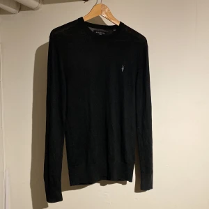 Svart stickad tröja från AllSaints - En svart, tunnstickad tröja från AllSaints med rund hals och diskret logga på bröstet. Tröjan har lång ärm och klassisk passform, perfekt för lager-på-lager. Enkel och stilren design som funkar till allt. Varan är använd max 2 gånger som helt ny. Hör av dig vid minsta fundering