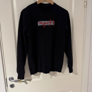Svart sweatshirt från Hugo Boss - Svart sweatshirt från Hugo Boss med broderad Hugo-logga i rött och grått framtill. Tröjan har rund hals, långa ärmar och ribbade muddar. Perfekt för en clean och stilren look. Pris ej fast kom med frågor
