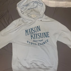 Ljusblå Maison Kitsuné hoodie - Säljer en ljusblå hoodie från Maison Kitsuné med stor tryckt logga i mörkblått framtill. Tröjan har huva med snörning och en klassisk känguruficka. Perfekt för dig som gillar streetwear och vill ha något stilrent men ändå trendigt. Pris går att diskuteras