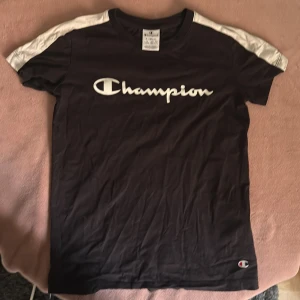 Svart Champion t-shirt med vita detaljer - Svart t-shirt från Champion i storlek XS med vita paneler på axlarna och stort Champion-tryck över bröstet. Klassisk rund hals och korta ärmar. Perfekt för en sportig och avslappnad stil.