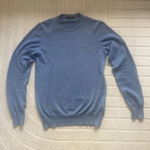 Ljusblåa merinoulltröja från J.Lindeberg - Säljer en stilren blå tröja från J.Lindeberg i mjuk merinoull. Tröjan har rund halsringning, diskret JL-logo på bröstet och ribbade muddar vid ärmslut och nederkant. Perfekt för lager-på-lager och snygg till jeans eller chinos.