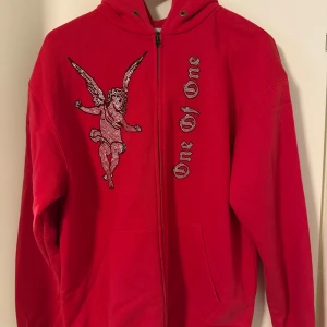 Röd hoodie One Of One med ängel - Säljer en röd hoodie från One Of One med dragkedja, huva och fickor. Framsidan har en stor strassutsmyckad ängel och texten 'One Of One' i bling. Perfekt statement-plagg för dig som vill sticka ut. 
