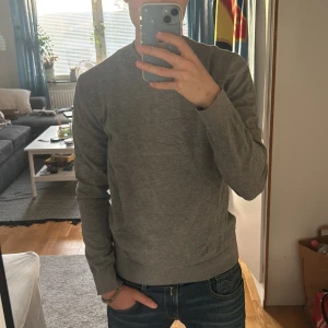 Grå slim fit tröja från H&M - Säljer en stilren grå tröja från H&M i slim fit-modell. Tröjan har rund halsringning och långa ärmar, perfekt för lager-på-lager. Tillverkad i mjukt material som känns skönt mot huden. Passar dig som gillar en enkel och clean look.