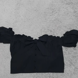 Svart offshoulder topp PrettyLittleThing - Svart croppad offshoulder topp från PrettyLittleThing med puffiga ärmar, knappar framtill och en stor rosett vid bysten. Toppen har volangdetaljer längs kanten och är perfekt för dig som vill sticka ut lite extra.