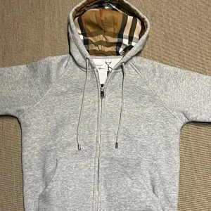 Grå Burberry hoodie med dragkedja - Snygg grå hoodie från Burberry med klassiskt rutigt foder i huvan och broderad logga på bröstet. Tröjan har dragkedja, snörning i huvan och är tillverkad i mjuk bomull. Perfekt för dig som vill ha en stilren och exklusiv look. Motsvarar storlek 158 CL