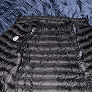Svart Moncler pufferväst  - Säljer en svart pufferjacka från Moncler i storlek L. Jackan har quiltad design, dragsko vid huvan och en innerficka med dragkedja. Materialet är glansigt och känns lyxigt, perfekt för kalla dagar. Klassisk Moncler-logga på bröstet. Storleken är L men passar även för folk som har M som storlek
