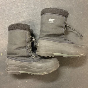 Svarta vinterstövlar från Sorel - Svarta vinterstövlar från Sorel med grov sula och snörning framtill. Ovandel i slitstark textil och gummiförstärkt nederdel. Fodrad insida och hög modell som håller värmen. Perfekta för kalla dagar och snö.