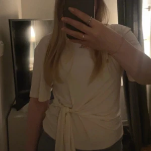 vit topp med knyt i midjan - Säljer en vit topp med kort ärm och rund halsringning. Toppen har en snygg knytning i midjan. Den har en liten sminkfläck på halsen, kan skicka bild på det. 
