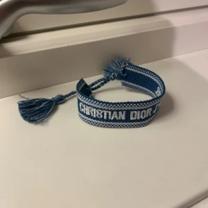 Blått dior armband - Snyggt blått armband från Christian Dior med vit broderad logga. Armbandet är vävt i tyg och har två tofsar i ändarna för att enkelt anpassa till din storlek.