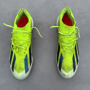 Adidas gula fotbollsskor med snörning - Säljer ett par Adidas fotbollsskor i neon-gult med svarta och röda detaljer. Skorna har snörning, platt sula med orange dobbar och ovandel i syntetmaterial. Insidan är röd och designen är sportig och modern, perfekt för dig som vill sticka ut på planen.