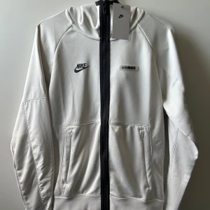 Nike Air Max hoodie med dragkedja - Nike Air Max hoodie i benvit färg med svart dragkedja och logga på bröstet. Tröjan har huva, två fickor framtill och är tillverkad i mjuk polyester. Perfekt för dig som gillar sportig och clean stil.
