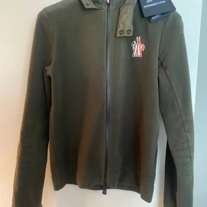 Olivgrön cardigan jacka från Moncler - Snygg olivgrön cardigan jacka från Moncler med dragkedja framtill och broderad logga på bröstet. Jackan har hög krage med knappdetaljer och Monclers signaturband vid ärmslut. Perfekt för dig som vill ha en stilren och exklusiv look.