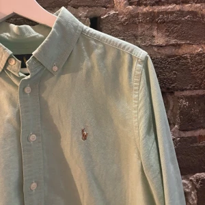 Mintgrön skjorta från Ralph Lauren - Snygg mintgrön skjorta från Ralph Lauren med klassisk button down-krage och broderad logga på bröstet. Skjortan har långa ärmar, vita knappar och är gjord i mjuk bomull. Perfekt för en clean och fräsch look.
