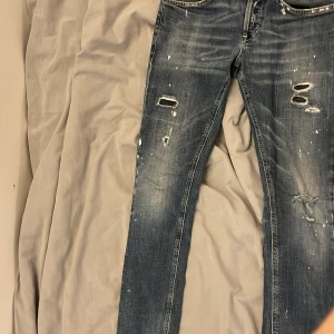 Blå slitna jeans från Dondup, stl 29 - Säljer ett par fett jeans från Dondup med snygga slitningar och på , samt vita färgstänk look. Modellen är smal och sitter snyggt på kroppen. Klassiska fem fickor och orangea detaljer på bakfickorna. Perfekt för dig som gillar trändiga kläder. Skriv gärna om du har frågor.🤗