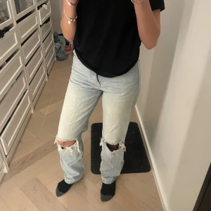 Ljusblå ripped jeans från  - Säljer ett par ljusblå jeans från ginatricot i storlek 34. Jeansen har raka ben, hög midja och slitna detaljer med stora hål vid knäna. Klassisk femficksmodell i bomull med dragkedja och knapp framtill. Perfekt för en avslappnad och trendig look.