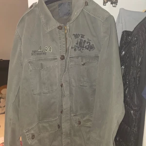 Grön overshirt jacka med tryck från Blend - Snygg militärgrön overshirt jacka från Blend med coola tryck och broderier både fram och bak. Jackan har fyra stora fickor framtill, knappar och dragkedja. Perfekt för dig som gillar streetstyle och vill sticka ut lite extra. Storlek passar bodde L och Xl