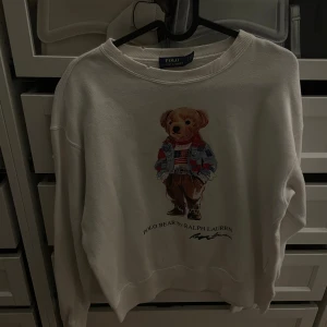 Vit sweatshirt Polo Bear Ralph Lauren - Vit sweatshirt från Polo Ralph Lauren med ikoniskt Polo Bear-tryck framtill. Tröjan har rund halsringning, ribbade muddar och är tillverkad i mjuk bomull. Perfekt för dig som vill ha en klassisk och trendig look med en lekfull touch.