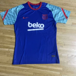 Blå FC Barcelona fotbollströja Nike - Snygg FC Barcelona fotbollströja från Nike i blått med röda detaljer och ljusblå mönstrade ärmar. Tryck med Beko på bröstet och ryggen samt klubbmärke på bröstet. Tillverkad i lätt och ventilerande polyester, perfekt för träning eller match.