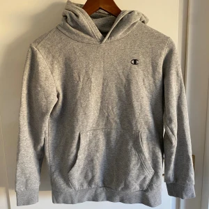 Grå hoodie från Champion - Säljer en klassisk grå hoodie från Champion med broderad logga på bröstet. Tröjan har huva, känguruficka och långa ärmar. Perfekt för chill dagar eller när du vill ha en enkel och stilren look.