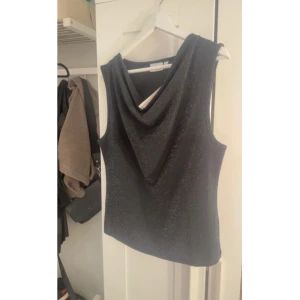 Glittrig svart ärmlös topp - Snygg svart ärmlös topp med glittrigt tyg och draperad halsringning. Perfekt för dig som vill sticka ut med en elegant och trendig look. Toppen har en smickrande passform och passar till många olika stilar.