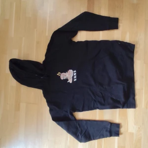 Svart hoodie från Vans med tryck - Svart hoodie från Vans med coolt tryck av en rosa blomma och logga på bröstet. Klassisk känguruficka framtill och huva. Perfekt för chill dagar eller när du vill ha en avslappnad stil.