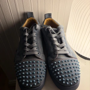 Blå Christian Louboutin sneakers med nitar - Säljer ett par blå sneakers från Christian Louboutin i mocka med ikoniska röda sulor och coola nitar över tån. Skorna har snörning och en rund tå, samt en snygg kontrast mellan den blå färgen och de ljusblå nitarna. Perfekta för dig som vill sticka ut. Pris kan diskuteras. Byten kan också diskuteras 