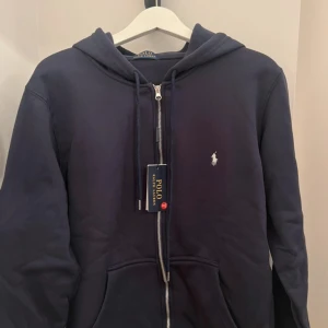  zip hoodie Polo Ralph Lauren - Snygg mörkblå hoodie från Polo Ralph Lauren med dragkedja, huva och klassisk logga broderad på bröstet. Tröjan har långa ärmar, dragsko i huvan och är tillverkad i mjuk bomull. Perfekt för en chill och stilren look.