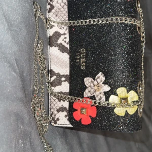 Glittrig axelväska från Guess med kedja - Snygg axelväska från Guess med glittrig svart framsida, ormskinnsmönster i vitt och grått samt dekorativa blommor i rött, gult och ormskinn. Väskan har silverfärgad kedjerem och logga på framsidan. Perfekt för dig som vill sticka ut med en unik accessoar.