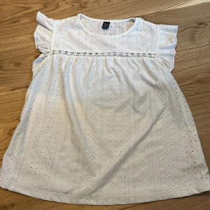 Vit blus med spets från SHEIN - Säljer en söt vit blus från SHEIN i storlek XS. Blusen har korta volangärmar, broderade detaljer och spetsband framtill. Materialet är bomull med ett genombrutet mönster som ger en fräsch och somrig vibe. Perfekt för dig som gillar romantisk stil! 