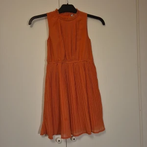 Ljust rostfärgad plisserad klänning från Friboo - Superfin ärmlös festklänning från Friboo med plisserad kjol och rynkad halsringning. Stängs med knapp i nacken och har fina veckdetaljer framtill.