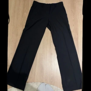 Svarta low waist kostymbyxor från Nelly, stl 36 - Svarta kostymbyxor från Nelly i avslappnad passform med låg midja. Byxorna har raka, vida ben och klassiska hällor för bälte. Perfekta för en clean och stilren look. 