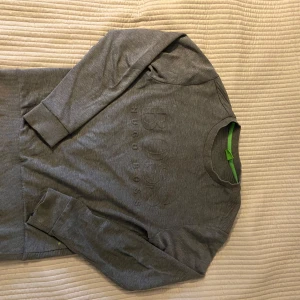 Grå sweatshirt från Hugo Boss - Snygg grå sweatshirt från Hugo Boss med broderad logga framtill. Tröjan har rund halsringning, ribbade muddar och en slim passform. Perfekt för dig som gillar stilrena och klassiska plagg med en modern touch.